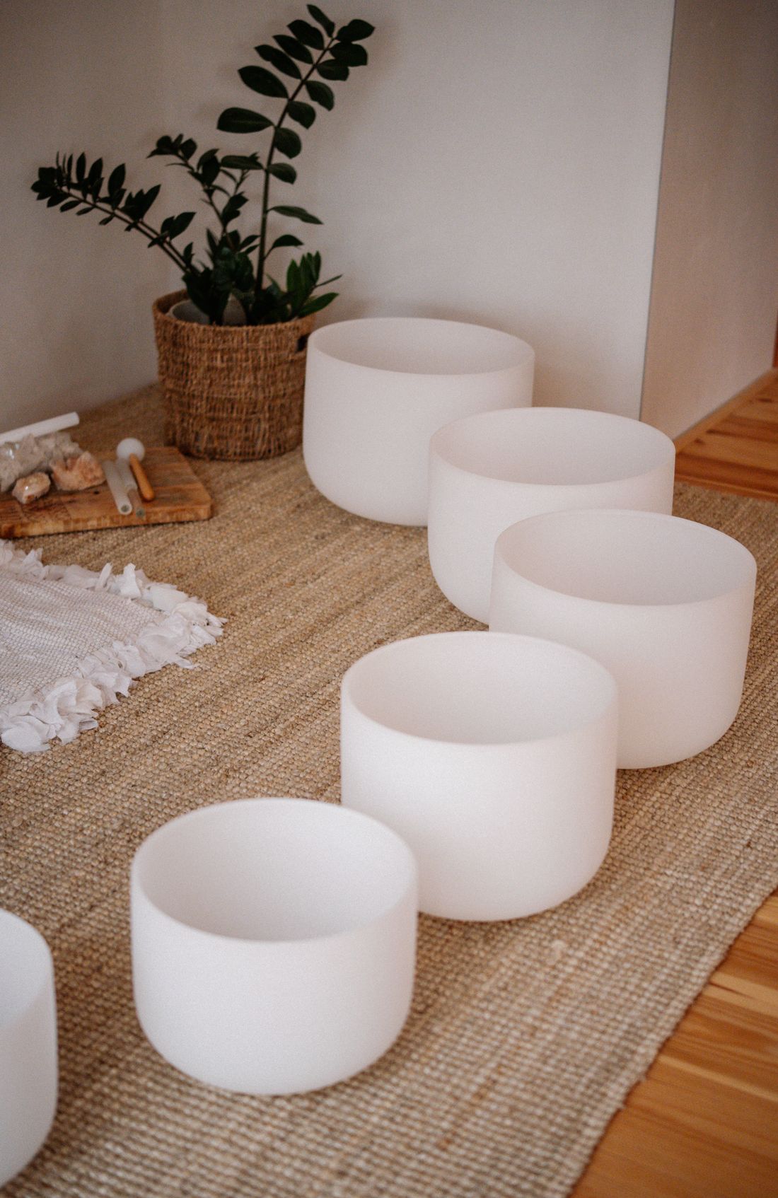 Six frosted crystal singing bowls arranged on a jute rug in a home, beside a potted plant in a woven basket.