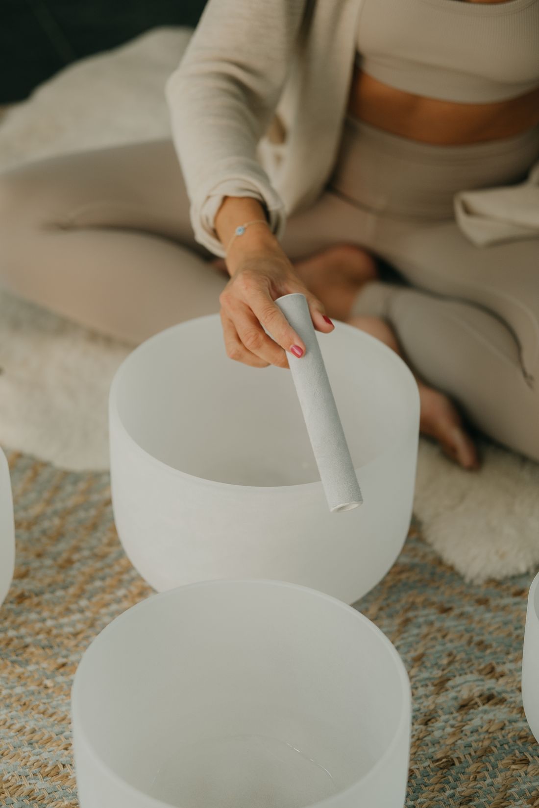 A seated person drawing a suede wand around the rim of a frosted crystal singing bowl on a woven rug.